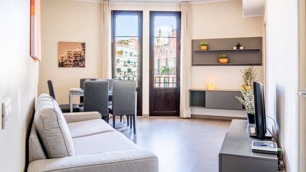 Ein modernes Wohnzimmer mit Sofa, Esstisch, Stühlen, einem Fernseher auf einem Schrank und einem Balkon mit Blick auf die Stadt.