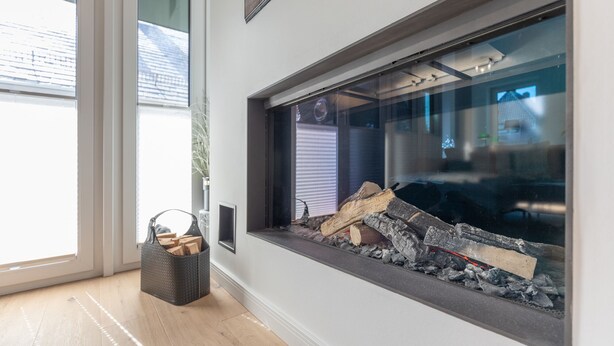 A modern interior with a large window, a fireplace with logs, and a basket of firewood on the floor.