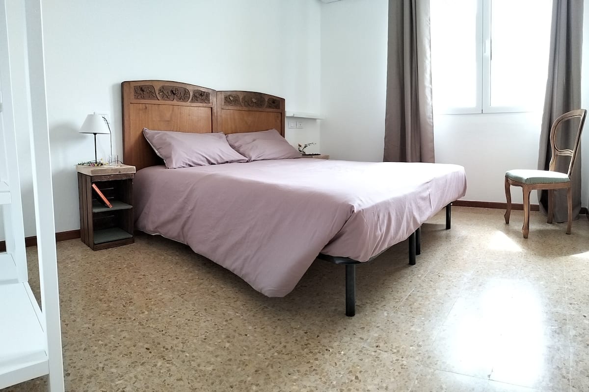 Een slaapkamer met een bed, een nachtkastje, een stoel en een raam met gordijnen.