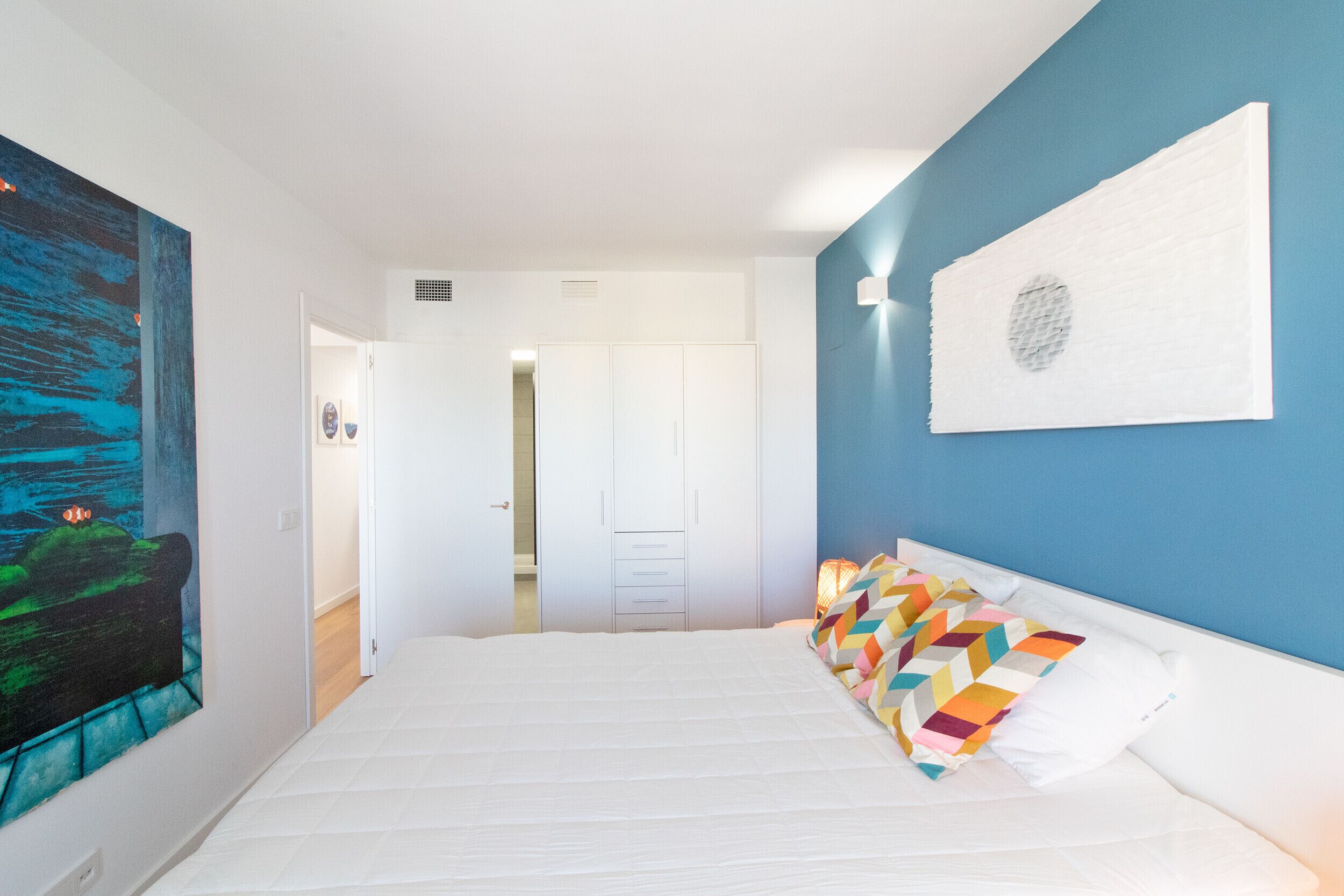 Een slaapkamer met een bed, kleurrijke kussens, een groot abstract schilderij en een blauwe accentmuur.