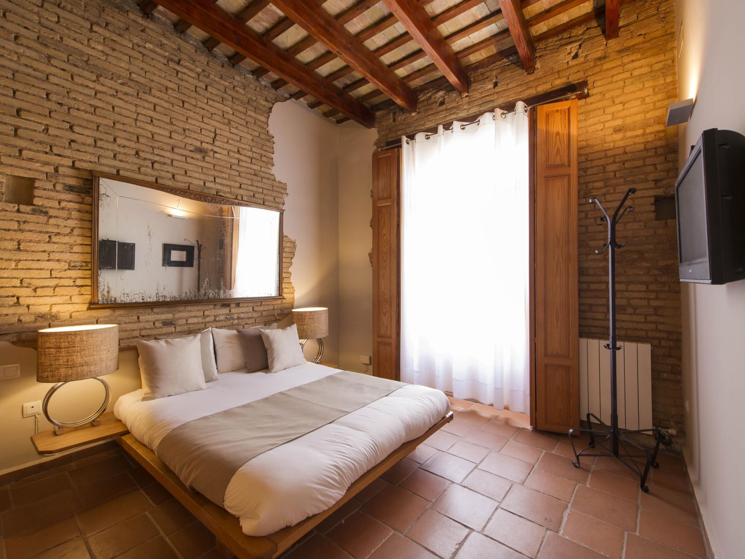Un dormitorio con pared de ladrillo, techo de madera, cama con ropa blanca, espejo, ventana con cortinas, radiador y televisor montado en la pared.