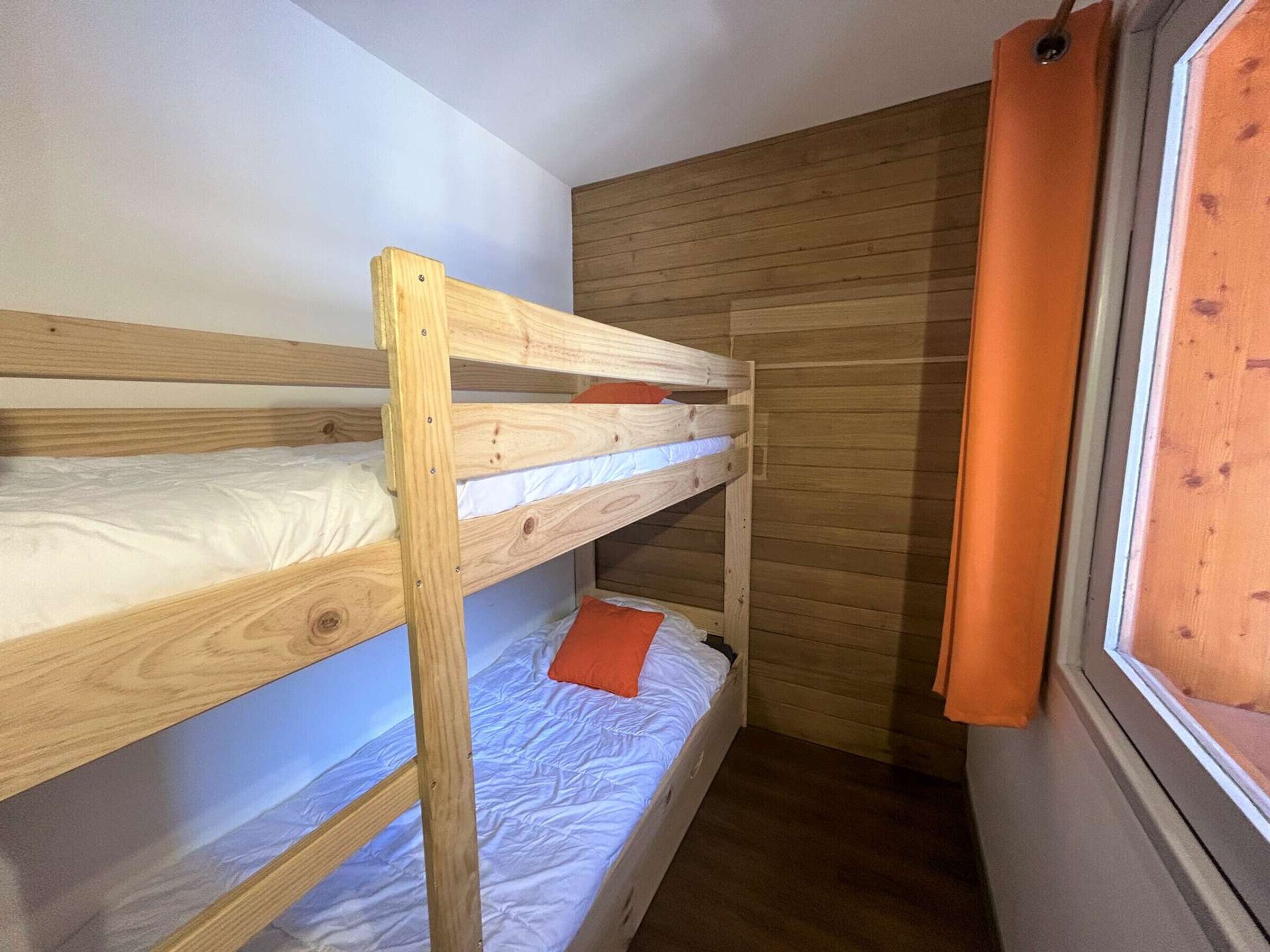 Une chambre avec des lits superposés, des murs en bois et une fenêtre ornée de rideaux orange.