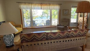 A cozy living room with a wicker sofa, a wooden coffee table, and a lamp. There are two windows with curtains, a chair, and a view of the outdoors. - Charming Lakeside Retreat: Cozy and Convenient on Chemong Lake (Lakefield)