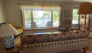 Una sala de estar acogedora con un sofá de mimbre, una mesa de centro de madera y una lámpara. Hay dos ventanas con cortinas, una silla y vistas al exterior.