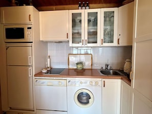 Cocina compacta con microondas, horno y lavadora. Hay armarios con puertas de vidrio y un lavabo.