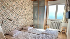 A bedroom with two beds, a patterned wallpaper, a radiator, a window with a view, and a television on the wall. - Charming 2-Room Apartment for 4 near Lifts - Alpe d'Huez (Huez)