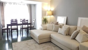 Ein modernes Wohnzimmer mit einem beigefarbenen Ecksofa, einem Esstisch mit Stühlen, einer Stehlampe und einem großen Fenster mit Vorhängen.