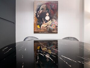 Uma zona de refeições moderna com uma mesa e cadeiras de mármore preto, um quadro pendurado na parede retratando uma mulher com joias, e uma obra de arte abstrata parcialmente visível.