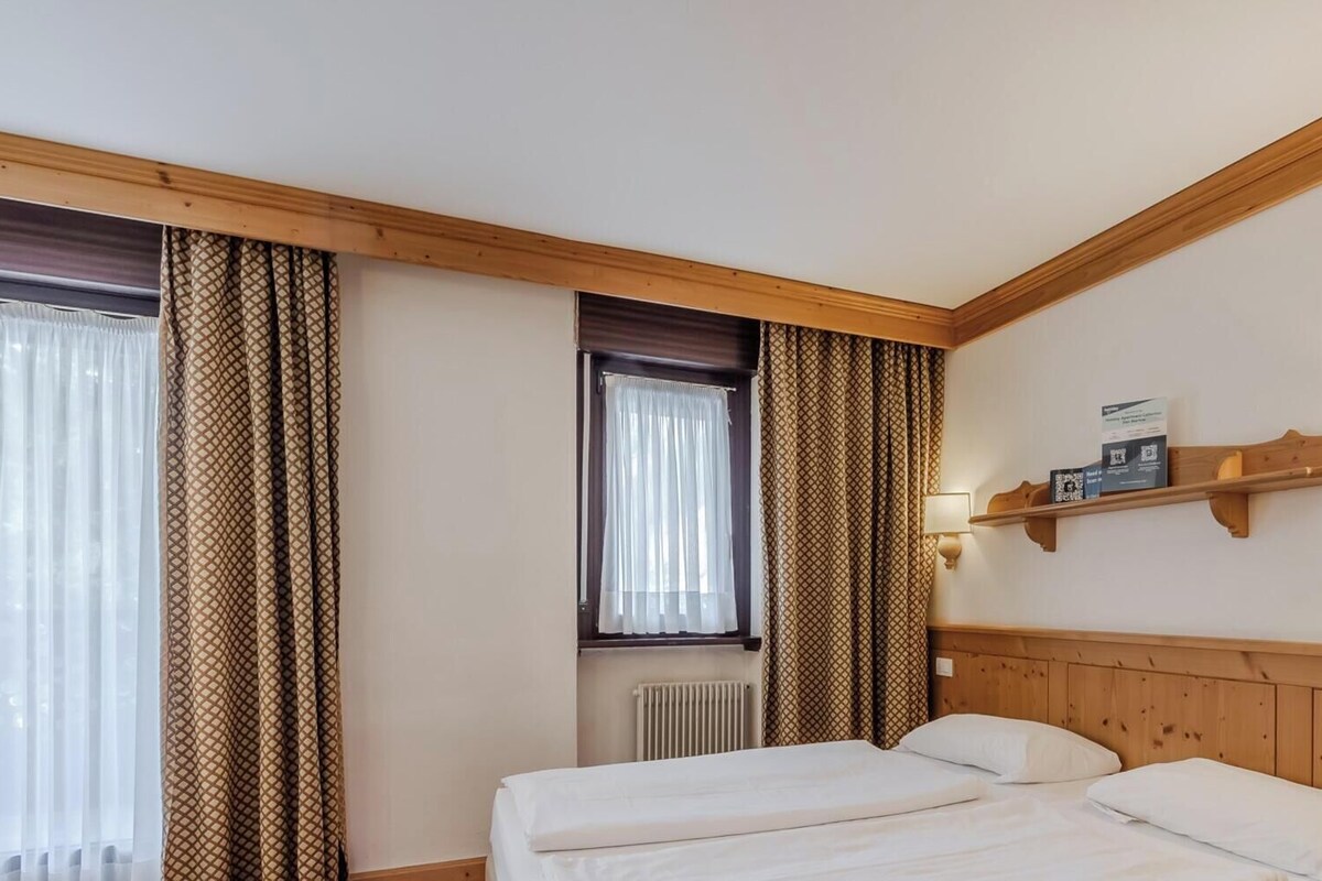 A hotel room with two beds, a window with curtains, a radiator, a shelf with a lamp, and a wall-mounted light.
