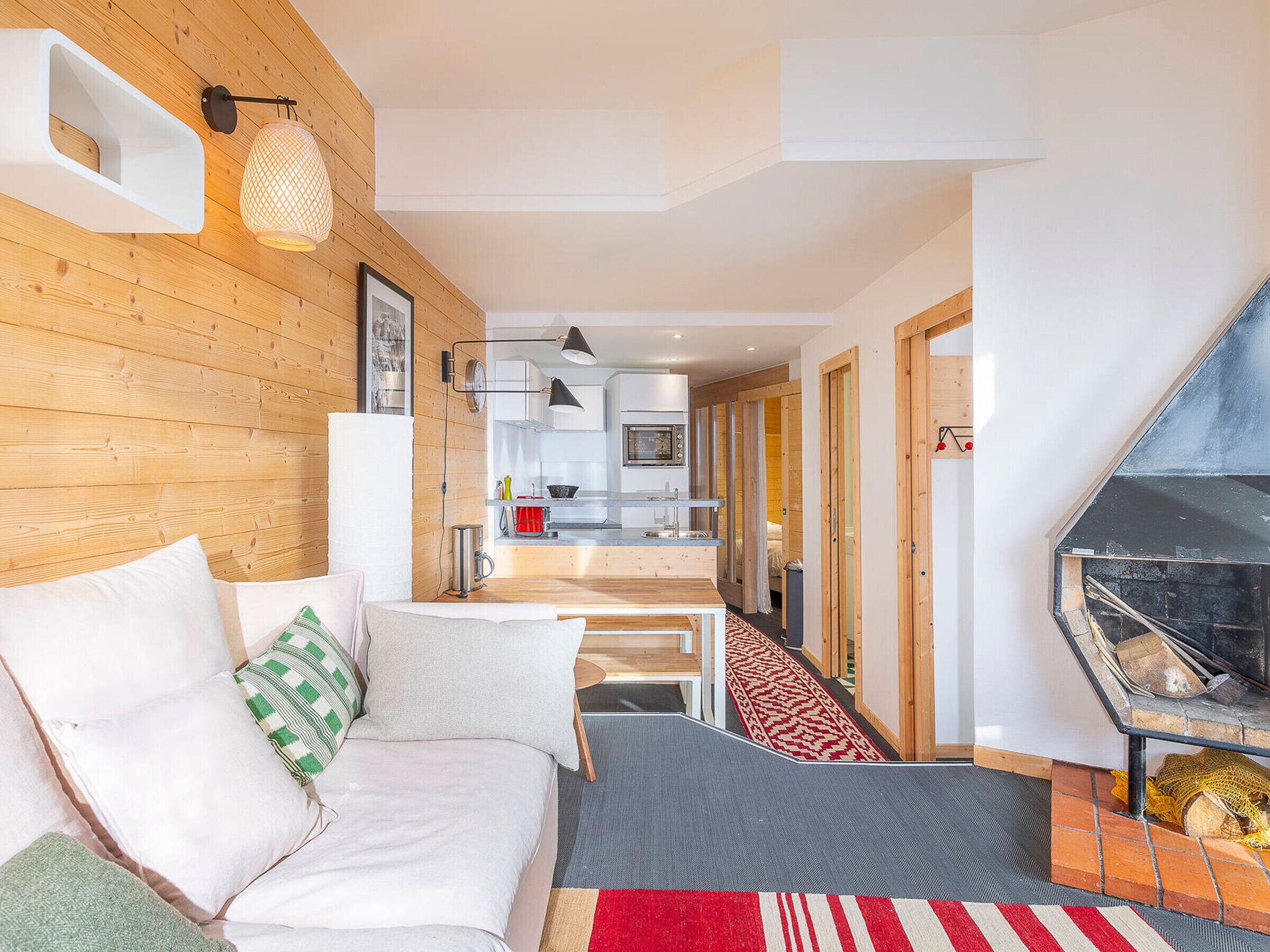 A modern living room with a wooden wall, a sofa with cushions, a fireplace, and a kitchen in the background.