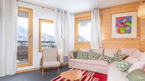 A cozy living room with a sofa, armchair, and a small table. There are large windows with curtains, a view of mountains, and a colorful abstract painting on the wall. - Cozy 2-room apartment in Avoriaz with fireplace, south-facing balcony (Avoriaz)