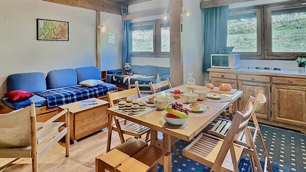A cozy room with a wooden ceiling, a blue sofa, a dining table with chairs, and a kitchen area with a window. - Spacious 2BR in Val d'Isère with Two Bathrooms (Val-d'Isère)