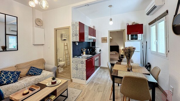 A modern living room with a sofa, coffee table, and dining area. There is a kitchen in the background with red cabinets and a dining table.