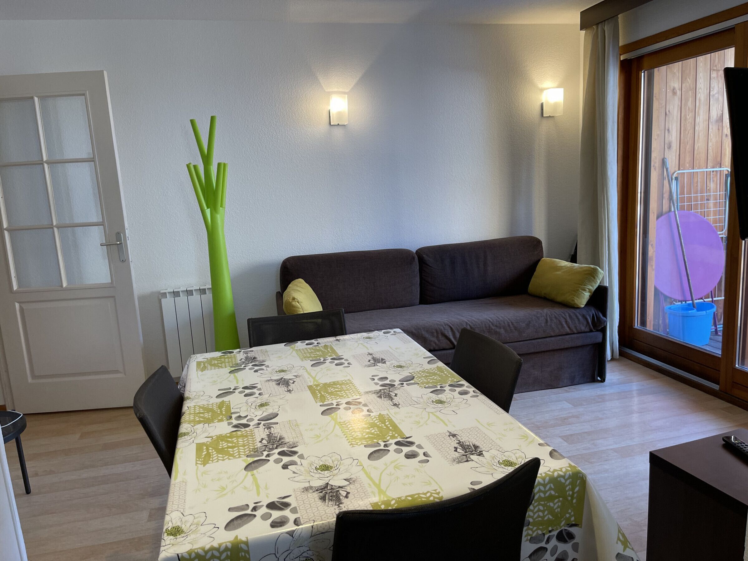 A room with a dining table, a sofa, and a window with a view of the outside.