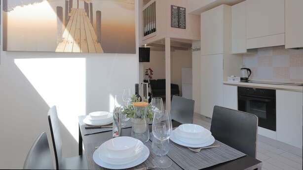 A dining area with a table set for four, a painting of a pier, and a kitchen in the background. - La Cà Fiera 23B. (Bologna)