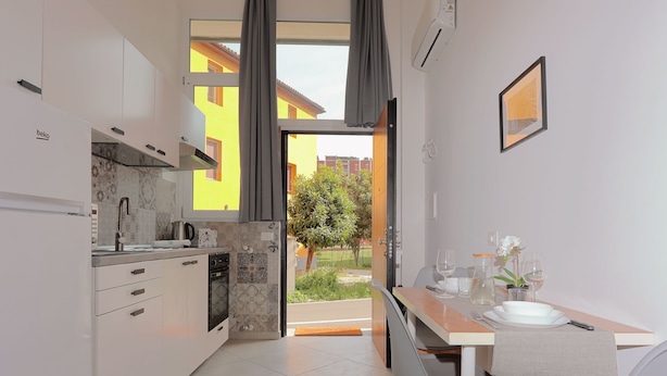 A modern kitchen with white cabinets, a dining area with a table and chairs, and a view of a yellow building through an open door. - La Cà Fiera 11E. (Bologna)