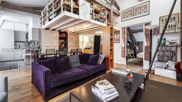 A modern living room with a purple sofa, a coffee table with books, and a loft area with shelves and artwork.