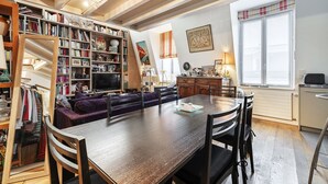 A cozy living room with a wooden dining table, purple sofa, and a bookshelf filled with books and decorative items.