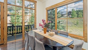 Een eetgedeelte met een houten tafel en stoelen, grote ramen met houten kozijnen en uitzicht op een woongebied met huizen en bomen.
