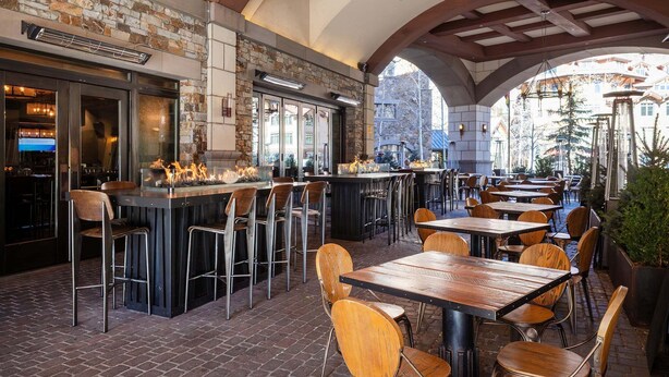 Een terras met bakstenen vloer, houten tafels en stoelen, een stenen muur en een bar met krukken.