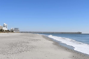 Una playa con un muelle largo que se adentra en el agua, un edificio de fachada blanca y un cielo azul despejado.
