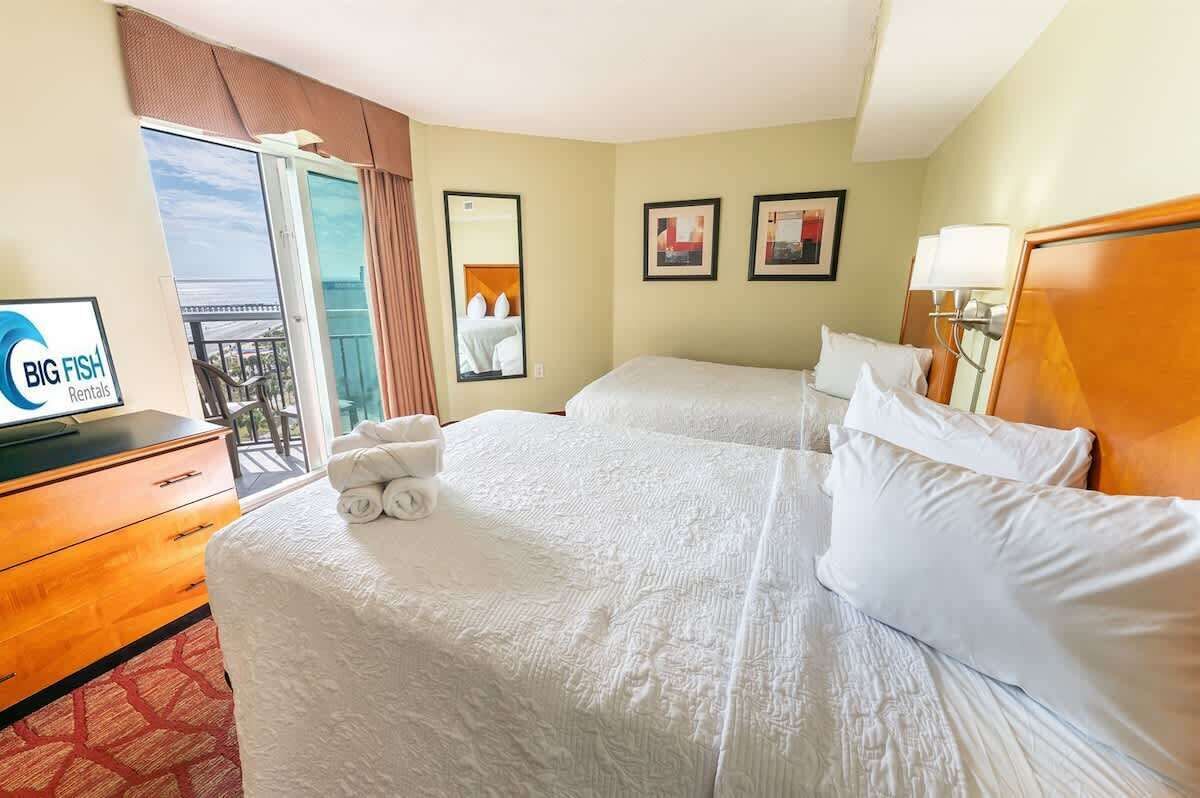 Une chambre d’hôtel avec deux lits, une commode en bois, un bureau avec un ordinateur, une fenêtre donnant sur le balcon et deux tableaux encadrés au mur.