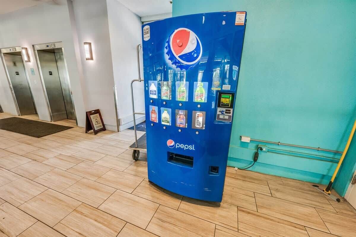 Eine blaue Pepsi-Getränkeautomat in einer Empfangshalle mit Fliesenboden, einer Tür und einer wandmontierten Steckdose.