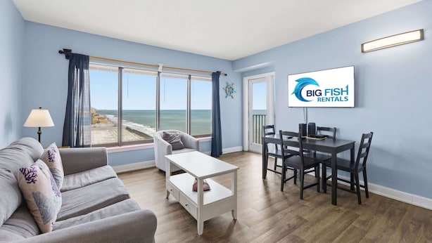 Ein Wohnzimmer mit Sofa, Couchtisch, Esstisch mit Stühlen, Fernseher und Blick auf den Strand.