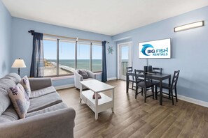 A living room with a sofa, a coffee table, a dining table with chairs, a TV, and a view of the beach. - New Palace Resort 2BR w/ Pools, High Floor w/Views (Myrtle Beach)