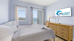 Ein Schlafzimmer mit Bett, Fernseher, Kommode und einem Balkon mit Vorhängen.