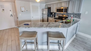 A modern kitchen with white cabinets, a granite countertop, stainless steel appliances, and two wooden stools. - Palace Resort 2BR, Pools & Ocean Views, 2nd Floor (Myrtle Beach)