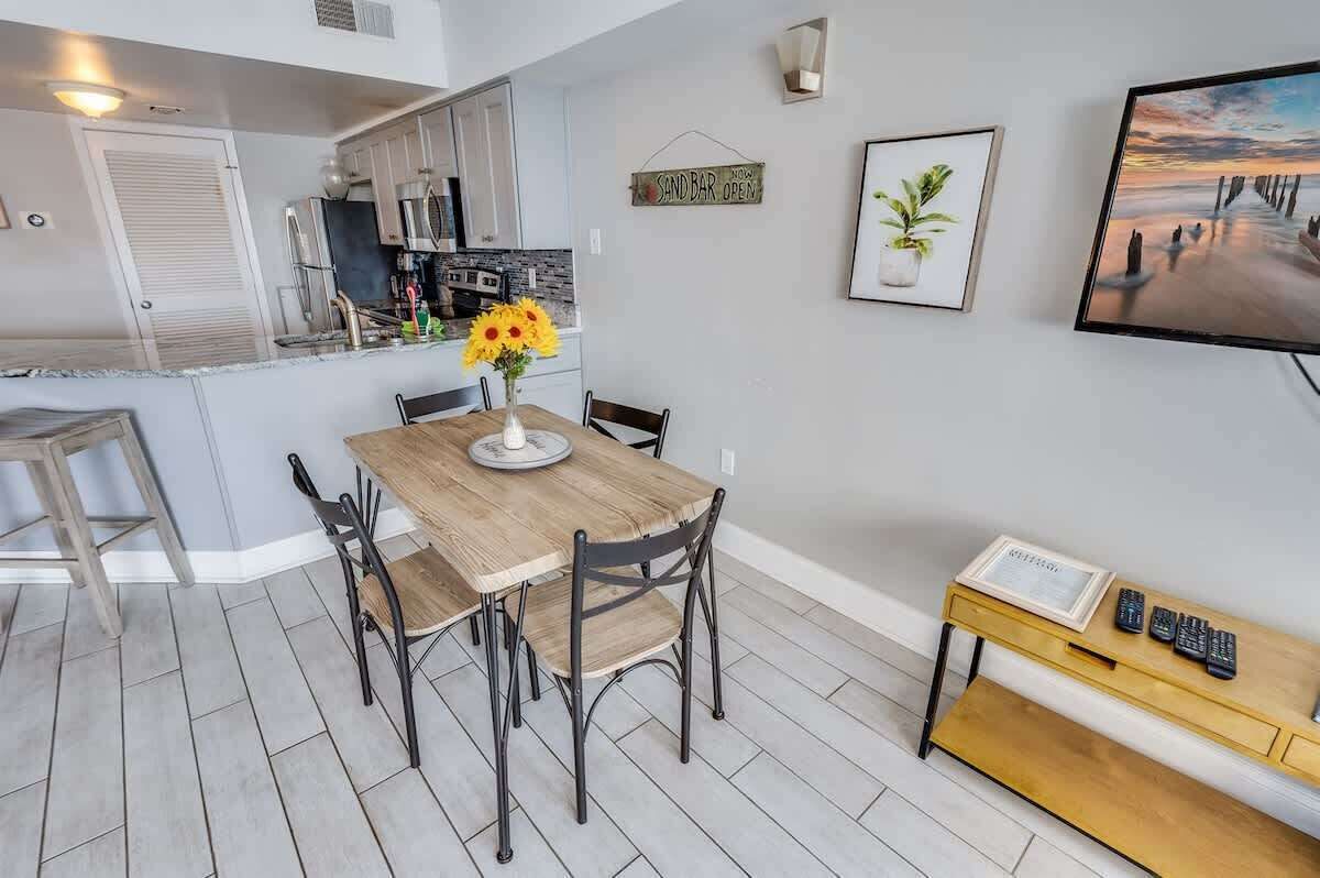 A modern kitchen with white cabinets, a wooden dining table with four chairs, a vase with sunflowers, and a TV on the wall.
