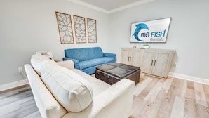 A living room with a white sofa, a blue sofa, a coffee table, and a cabinet. There is a sign that says "WIFI" and a TV screen with "BIG FISH Rentals" on it.