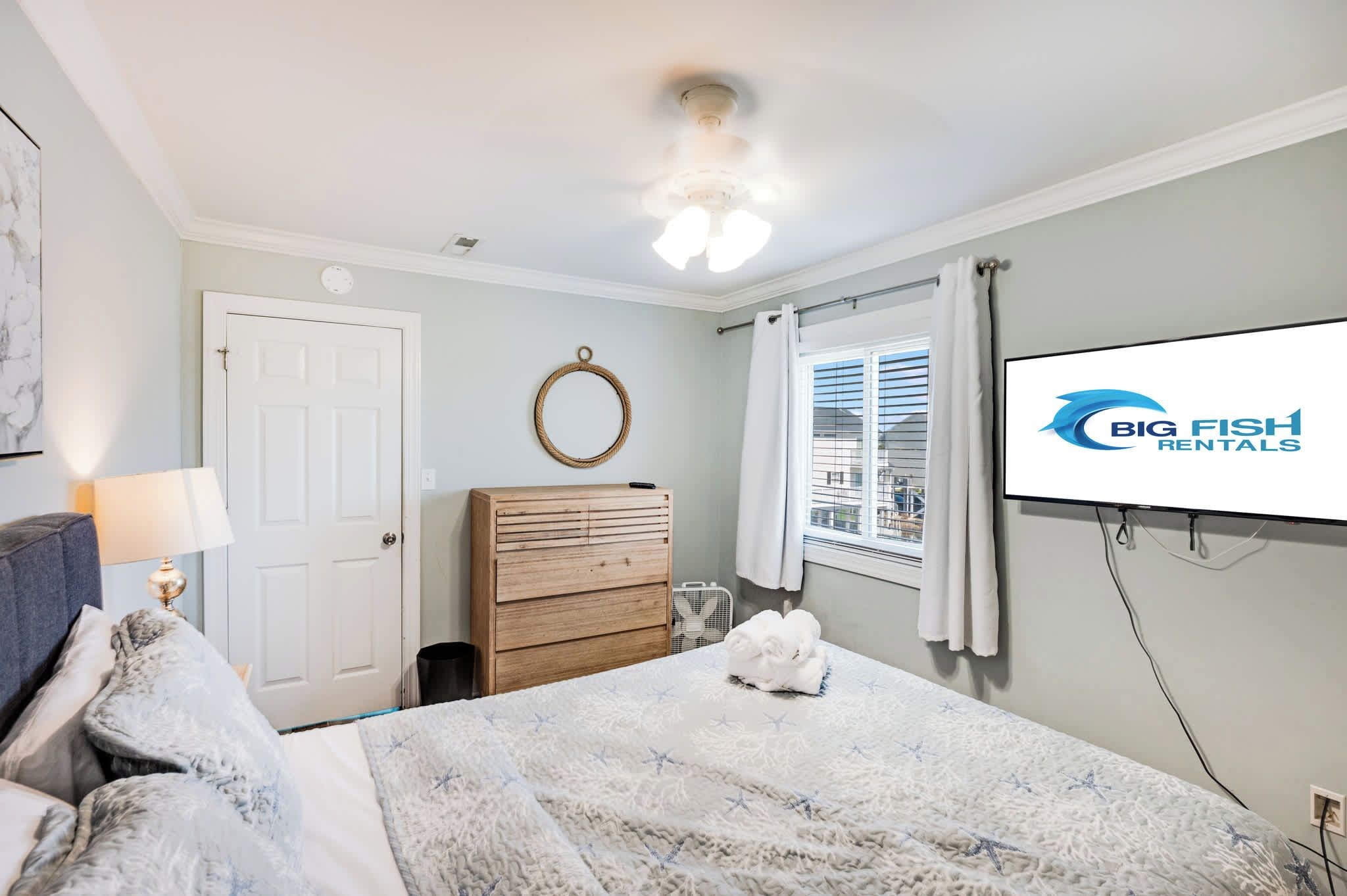 Quarto com cama, cômoda, TV na parede, ventilador de teto e janela com persianas.