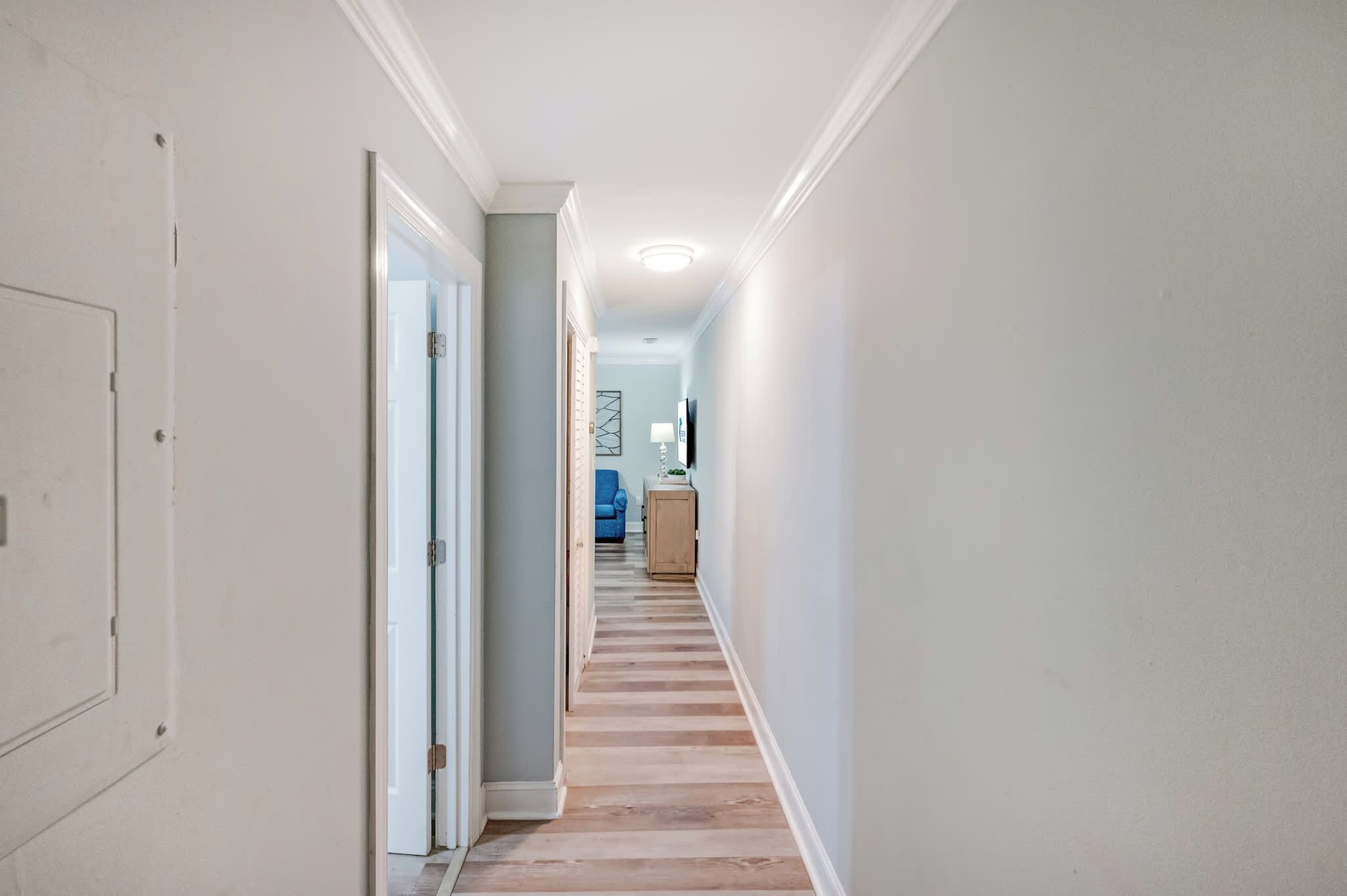 A hallway with a wooden floor, a door on the left, and a blue chair in the distance.