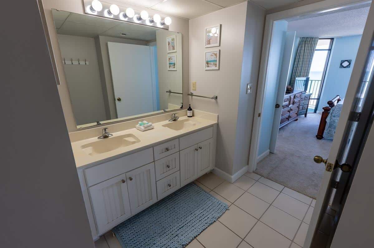 A bathroom with a double sink vanity, a large mirror with lights, and a door leading to a bedroom with a view.
