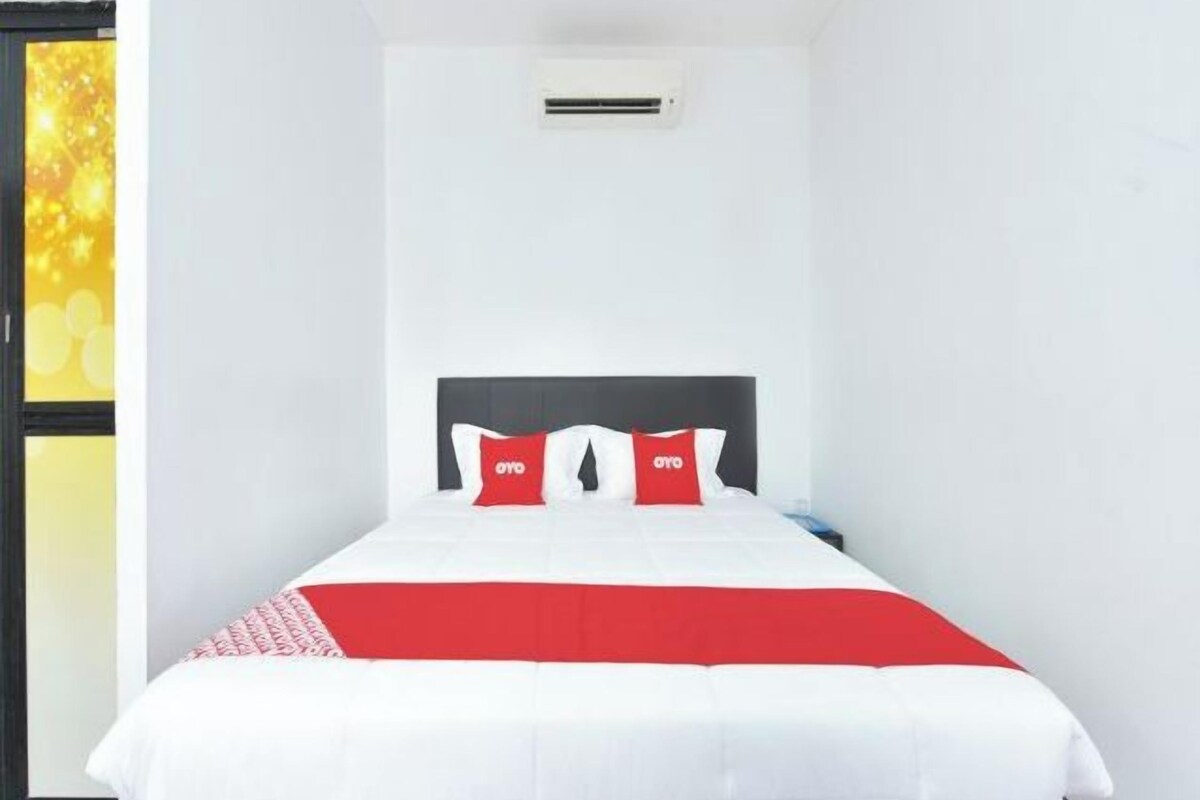 A hotel room with a bed, a red and white bedspread, a black headboard, and a small air conditioning unit on the wall.
