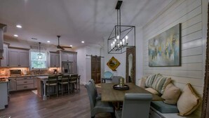 Una cocina moderna con armarios blancos, un comedor con mesa y sillas de madera, y una sala de estar con sofá y cojines.