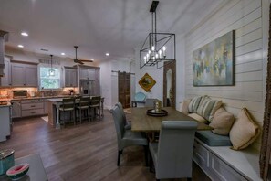 Eine moderne Küche mit weißen Schränken, ein Essbereich mit Holztisch und -stühlen sowie ein Wohnbereich mit Sofa und Kissen.