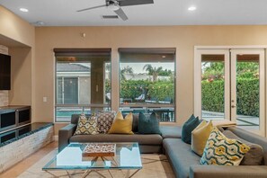 A modern living room with a glass coffee table, a sofa set, and a large window offering a view of a landscaped garden.