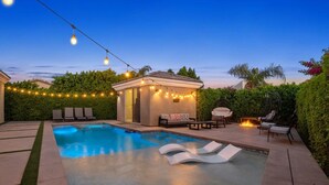 Una piscina con sillas de descanso, un pequeño pabellón con luces colgantes y una zona con fogata.
