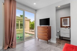 Un dormitorio moderno con puerta de vidrio corredera que da a una zona de piscina, un aparador de madera, un televisor y una mesita de noche.