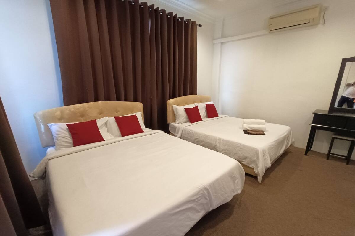 Habitación de hotel con dos camas, cada una con ropa blanca y almohadas rojas, cabeceros de madera, un escritorio con espejo y cortinas marrones.