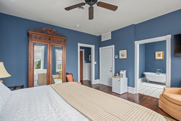 A bedroom with a large bed, a wooden wardrobe, a ceiling fan, a white door, a white dresser, a white bathtub, and a television on the wall.