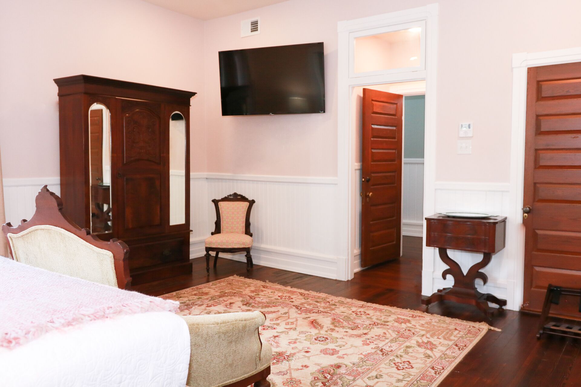 Un dormitorio con cama, una silla, un armario de madera, un televisor y una puerta que da a otra habitación.