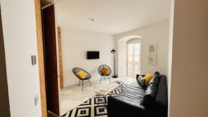 Sala de estar com sofá de couro preto, duas cadeiras de arame preto com almofadas amarelas, mesa de centro de madeira e tapete estampado.