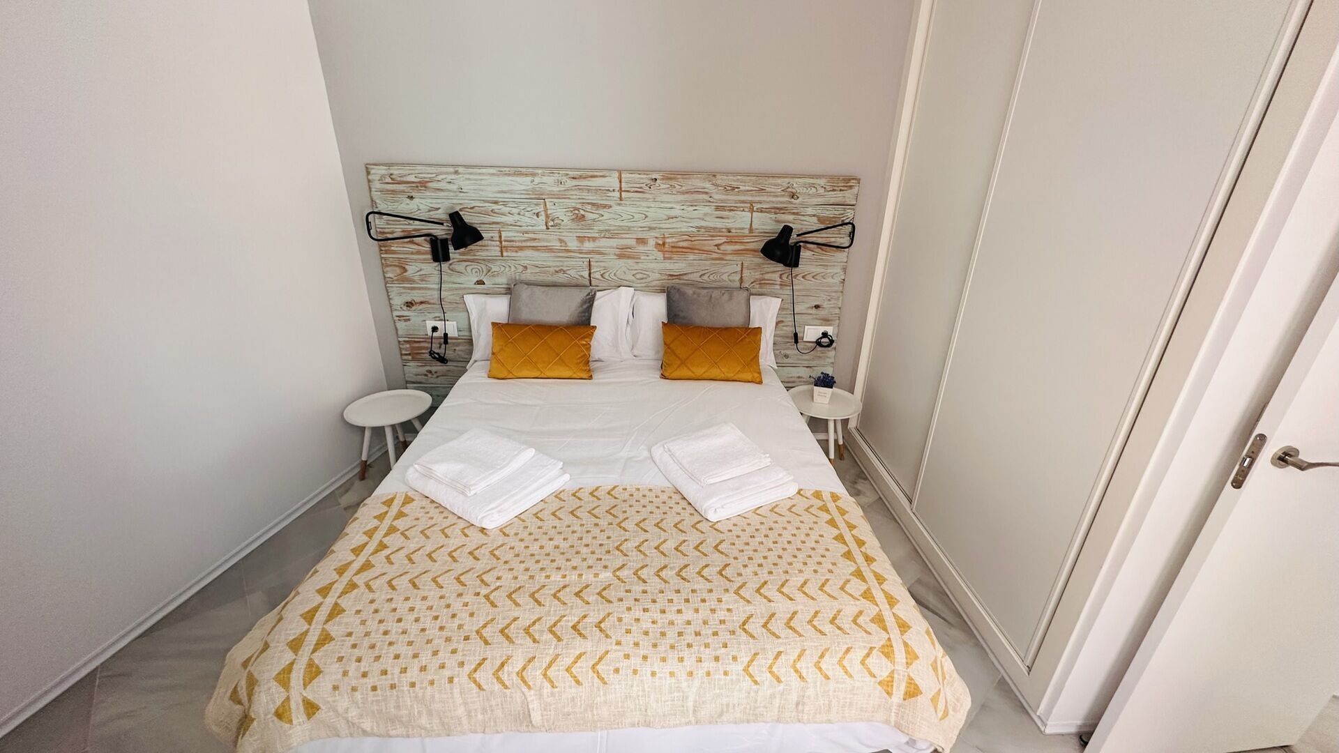 Una cama bien tendida con sábanas blancas y dos toallas dobladas, un cabecero de madera con acabado claro y dos lámparas de noche.