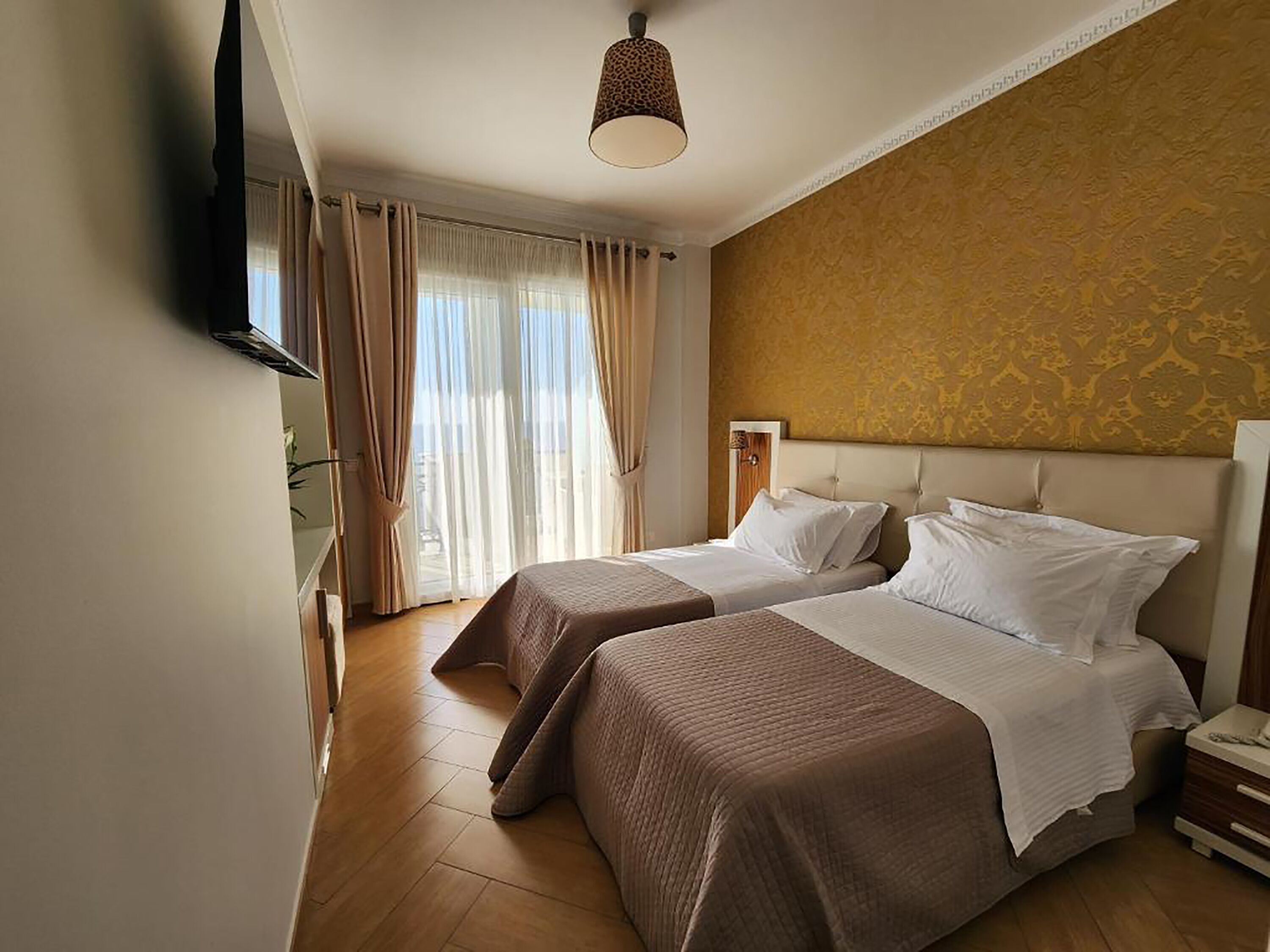 Ein Hotelzimmer mit zwei Betten, einem großen Fenster mit Vorhängen, einer Wand mit ornamentalem Wandpapier und einer Deckenleuchte.