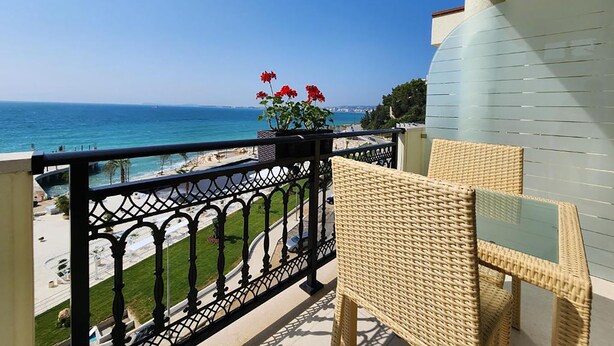 Un balcón con una silla de mimbre, una planta en maceta con flores rojas y vistas a la playa y al océano.