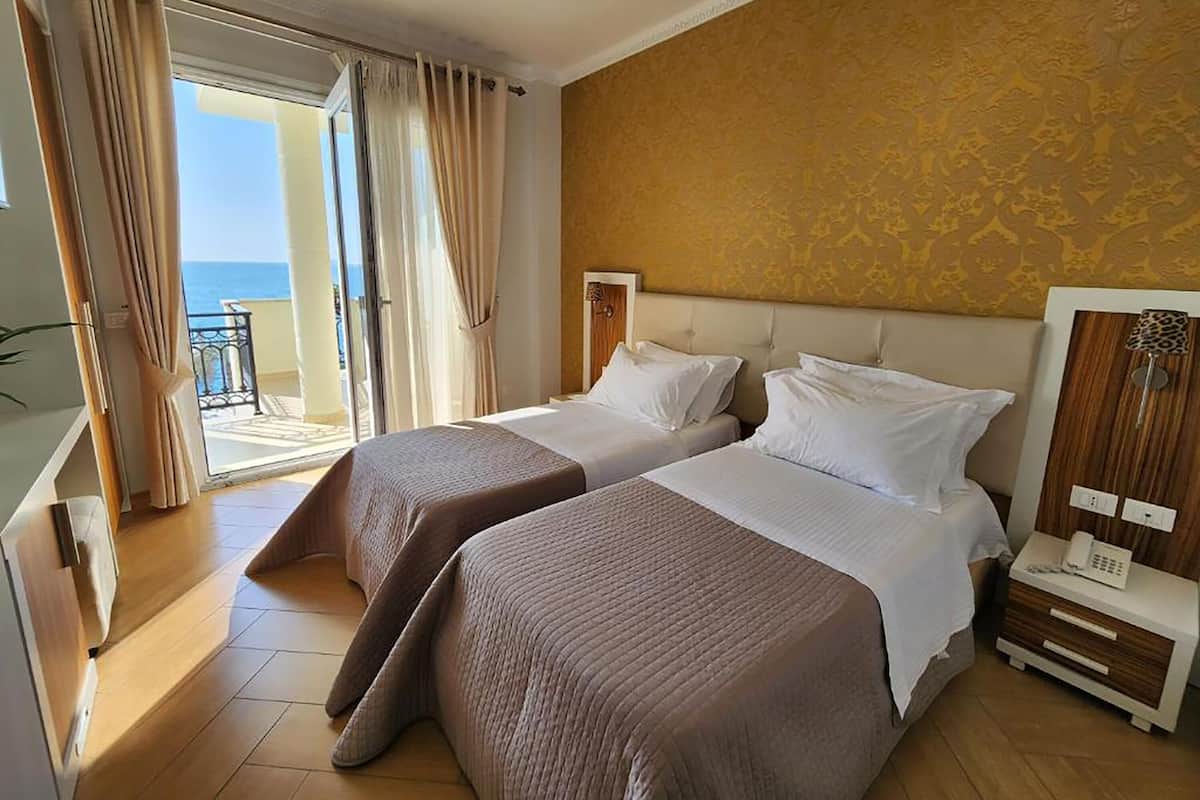 A hotel room with two beds, a balcony with curtains, a wooden floor, and a patterned wall.